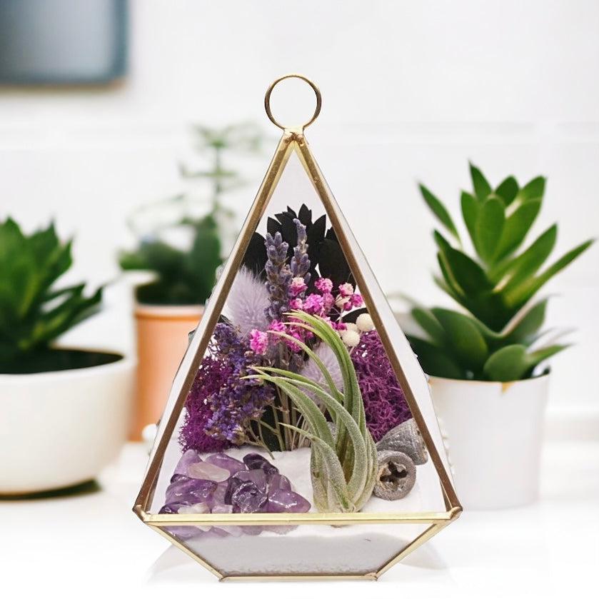 Gold-rimmed glass victorian terrarium with a dried flower bouquet with purple accents, sand, wood, moss, amethyst crystals and an airplant