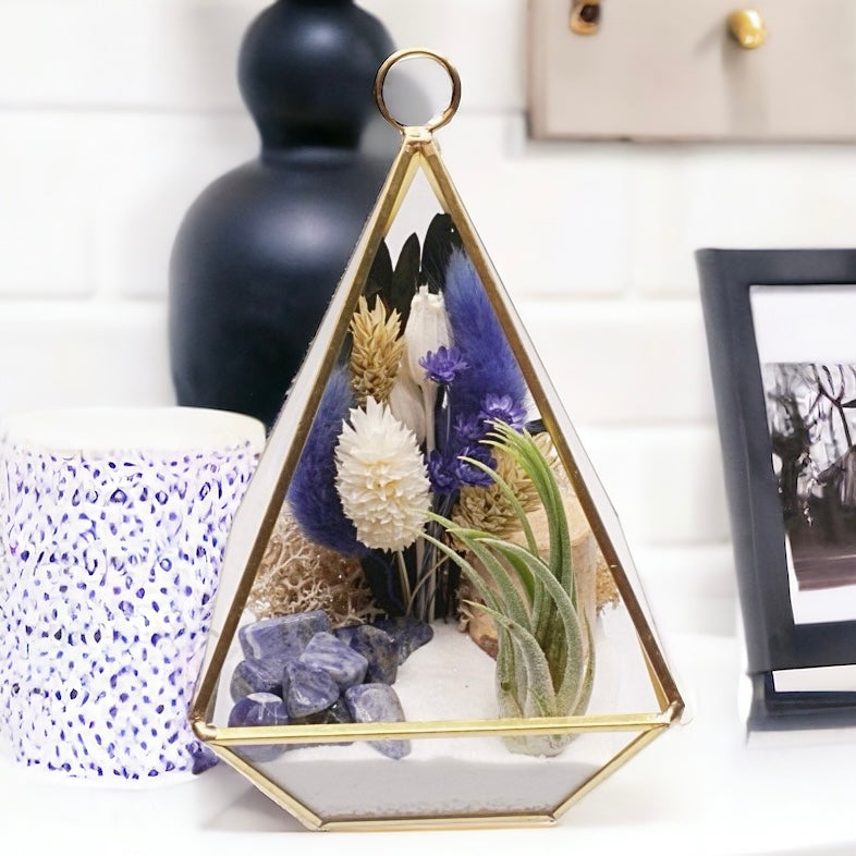 Gold-rimmed glass victorian terrarium filled with dried flowers with blue accents, polished sodalite crystals, sand, moss, wood and an airplant