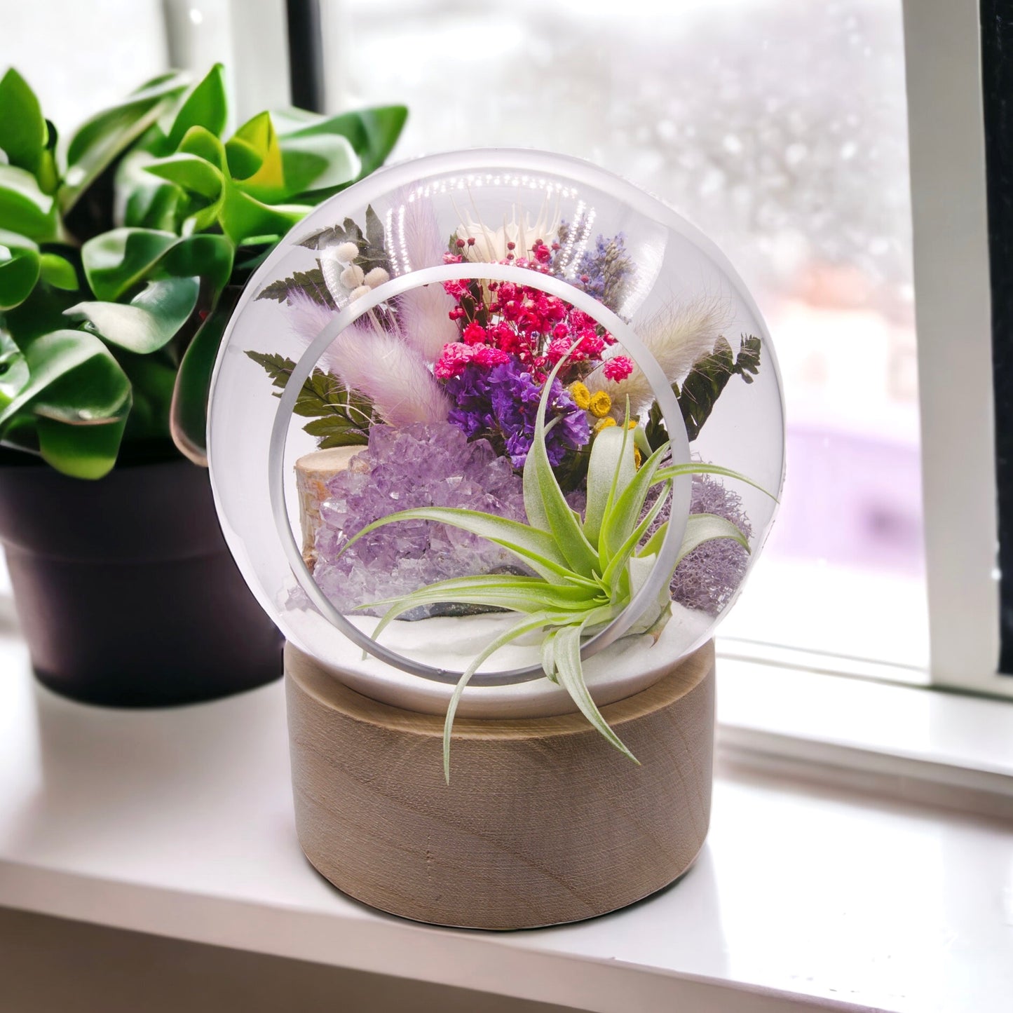 Glass bubble terrarium on a wooden base with dried flowers, sand, moss, wood and an airplant along with a raw amethyst crystal