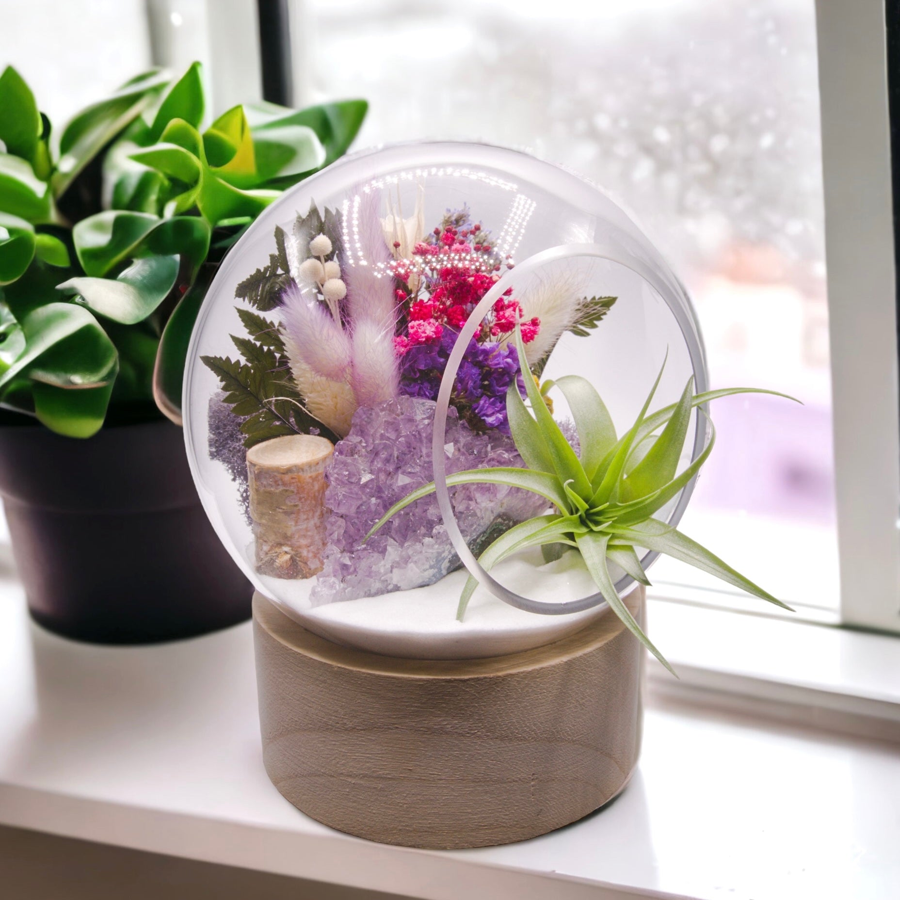 Glass bubble terrarium on a wooden base with dried flowers, sand, moss, wood and an airplant along with a raw amethyst crystal