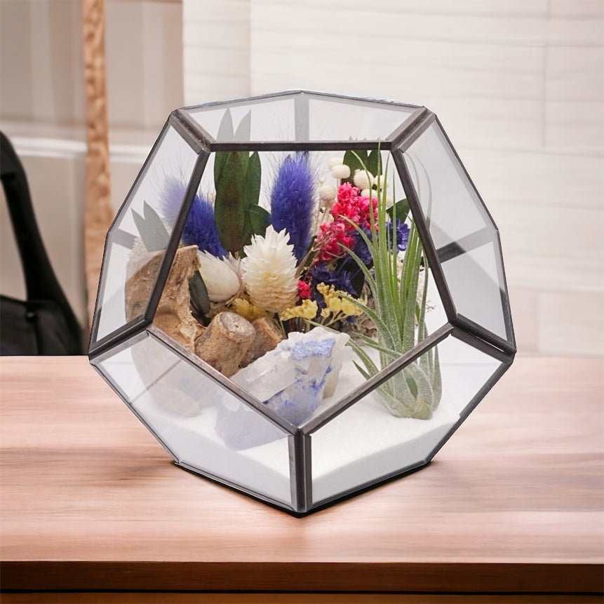 Black-rimmed glass geometric terrarium with an airplant, blue coloured dried flowers, sand, moss, wood and a blue quartz crystal