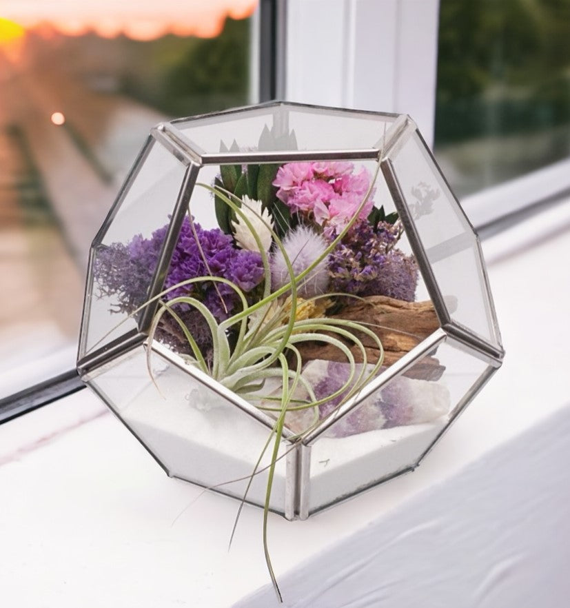 Black-rimmed glass victorian terrarium with a dried flower bouquet with purple accents, an amethyst chevron crystal, airplant, sand, moss and wood