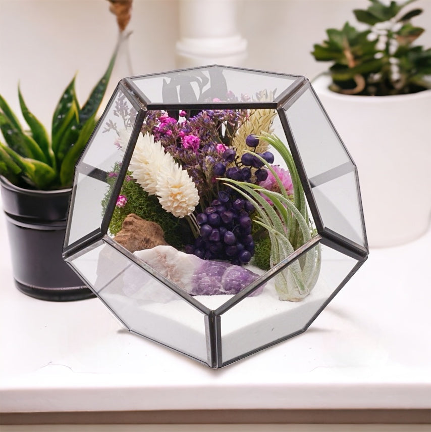 Black-rimmed glass victorian terrarium with a dried flower bouquet with purple accents, an amethyst chevron crystal, airplant, sand, moss and wood