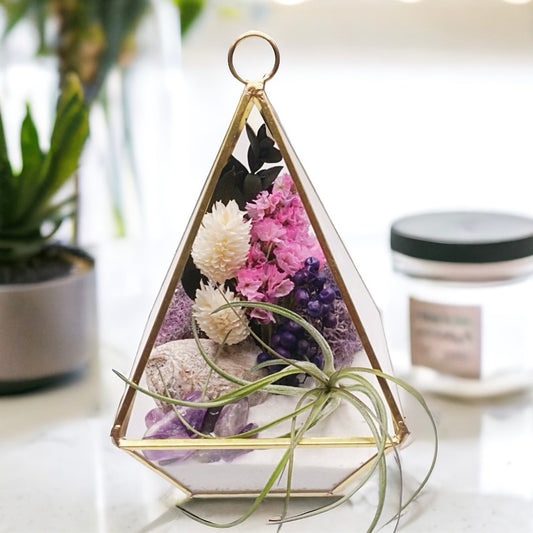 Gold-rimmed glass victorian terrarium with a dried flower bouquet with purple accents, sand, wood, moss, amethyst crystals and an airplant