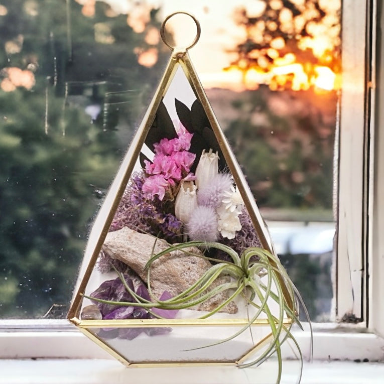 Gold-rimmed glass victorian terrarium with a dried flower bouquet with purple accents, sand, wood, moss, amethyst crystals and an airplant