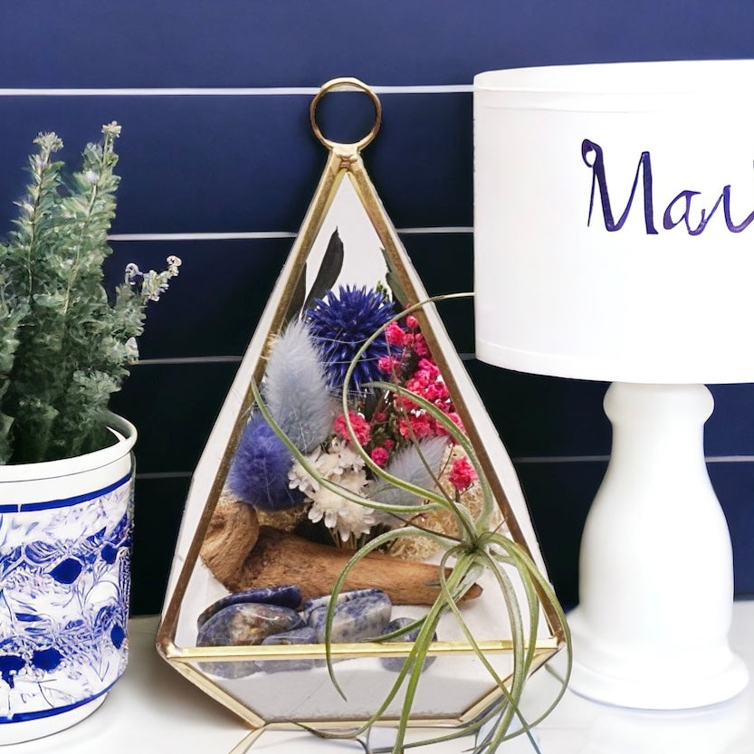 Gold-rimmed glass victorian terrarium filled with dried flowers with blue accents, polished sodalite crystals, sand, moss, wood and an airplant