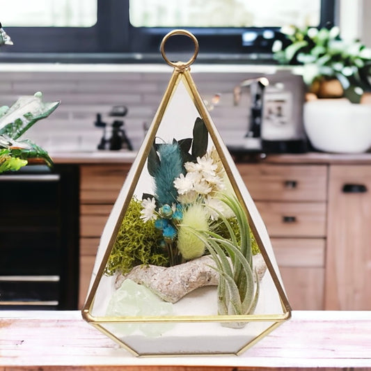 Gold-rimmed victorian glass terrarium filled with dried flowers with green accents, green calcite crystals, sand, moss, wood and an airplant