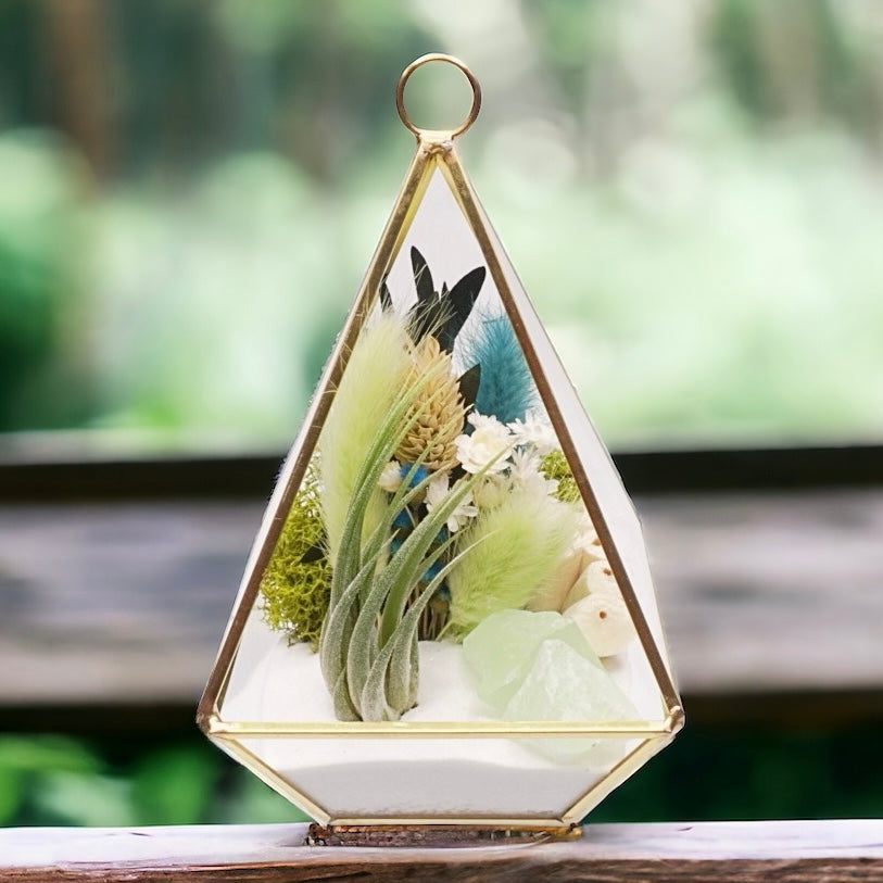 Gold-rimmed victorian glass terrarium filled with dried flowers with green accents, green calcite crystals, sand, moss, wood and an airplant
