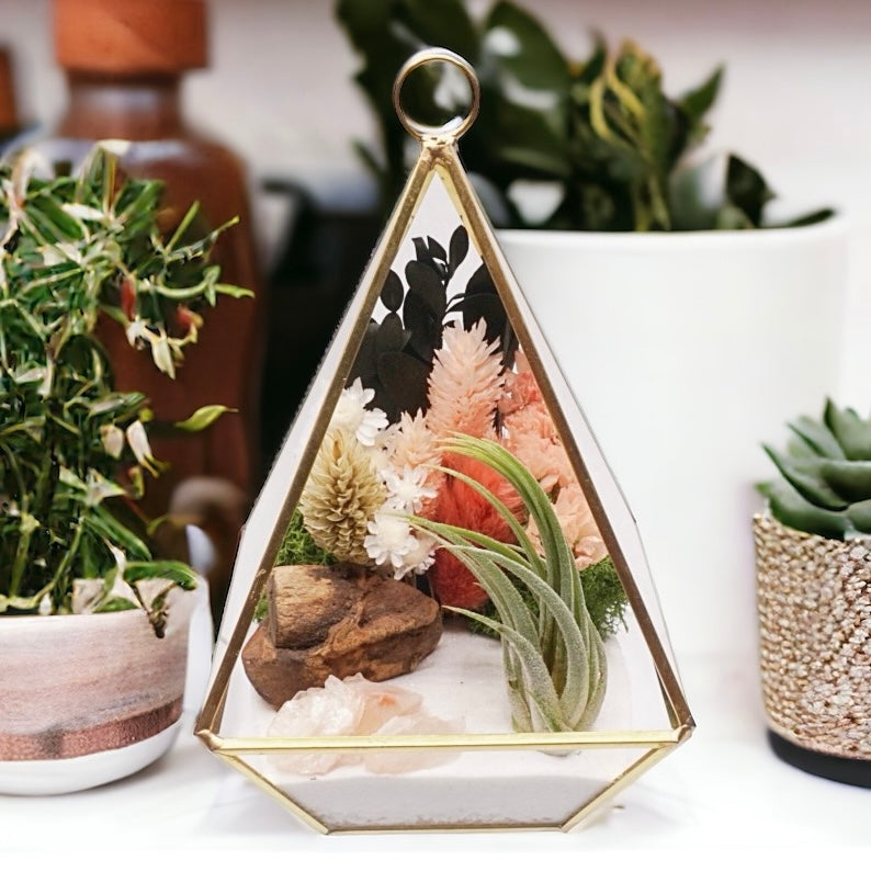 Gold-rimmed glass victorian terrarium filled with dried flowers with peach/terracotta accents, rose quartz crystals, sand, moss, wood and an airplant