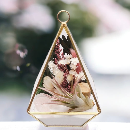 Gold-rimmed glass victorian terrarium filled with dried flowers with pink accents, rose quartz crystals, sand, moss, wood and an airplant