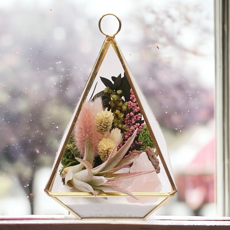 Gold-rimmed glass victorian terrarium filled with dried flowers with pink accents, rose quartz crystals, sand, moss, wood and an airplant