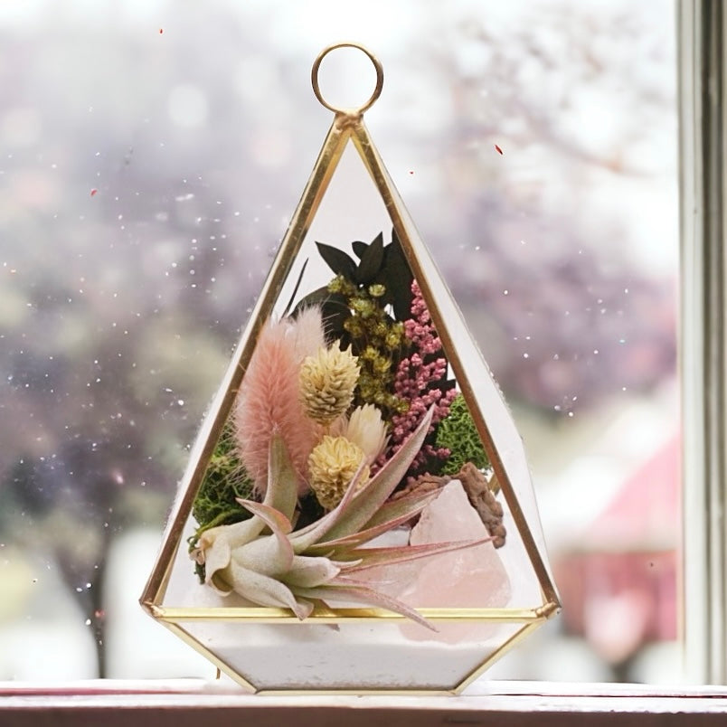 Gold-rimmed glass victorian terrarium filled with dried flowers with pink accents, rose quartz crystals, sand, moss, wood and an airplant