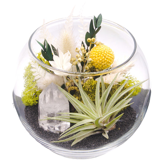 A glass bowl airplant terrarium with black sand, a bouquet of dried flowers, green moss, a dried mushroom and a crystal quartz tower.